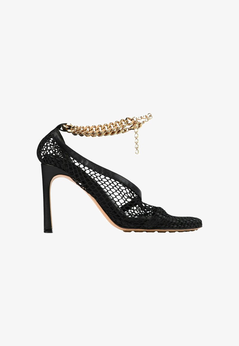 Chaussure à talon haut noire avec un design en mesh et une bride de cheville chaîne dorée, avec un talon fin et un bout pointu.