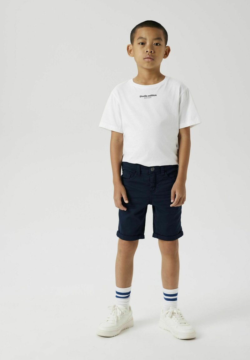 T-shirt blanche à manches courtes avec le texte "édition studio", associée à un short bleu marine et des baskets blanches. Chaussettes rayées horizontales. Design simple.