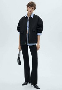 Black oversized jacket with a zipper, blue striped shirt underneath, black flared trousers, and pointed black shoes; short hair and handbag.