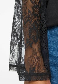 Svart kimono i blonder med blomsterdesign og transparent tekstur, med åpne ermer og glatt finish over en mørk topp og blå denim.