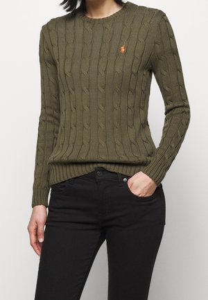 Femme portant un pull en maille torsadée vert olive avec un logo orange sur la poitrine et un jean noir, main dans la poche, sur un fond uni.