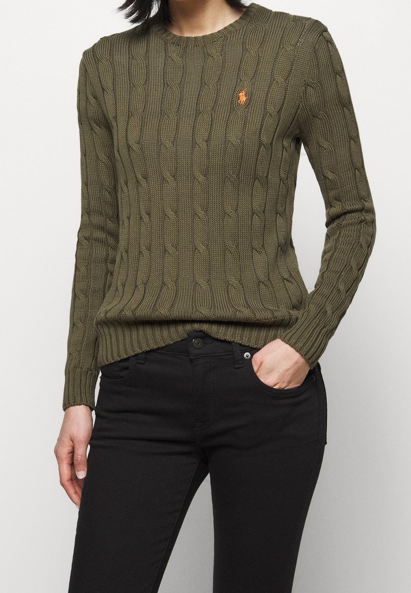 Femme portant un pull en maille torsadée vert olive avec un logo orange sur la poitrine et un jean noir, main dans la poche, sur un fond uni.