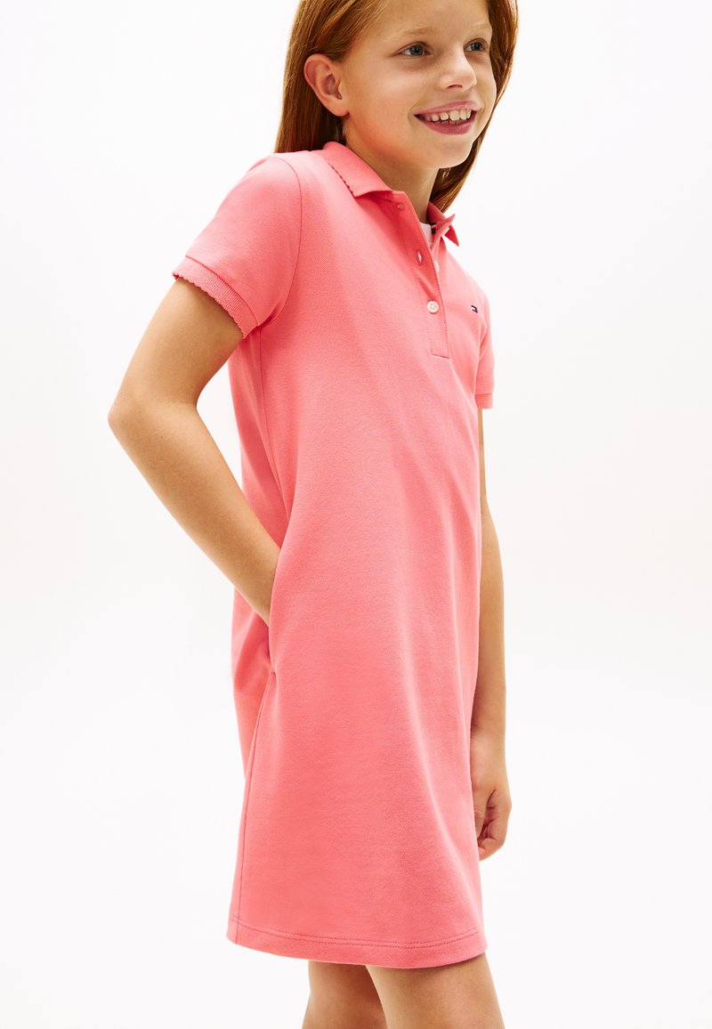 Νεαρή κοπέλα που χαμογελά, φοράει ροζ κοραλλί κοντομάνικο πόλο φόρεμα με το αριστερό χέρι στην τσέπη, στέκεται μπροστά σε λευκό φόντο.