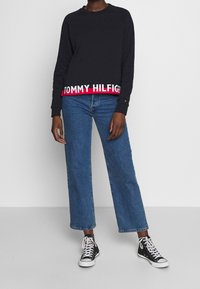 Felpa nera con logo "TOMMY HILFIGER" rosso e bianco sull'orlo, abbinata a jeans in denim blue dritti e sneaker alte nere.