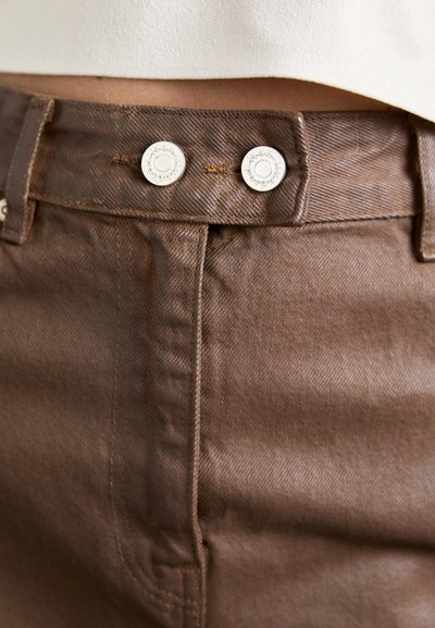 Brune bomulds-twill bukser med et nærbillede af en knappet talje, med to sølvmetal knapper og teksturerede stofdetaljer.