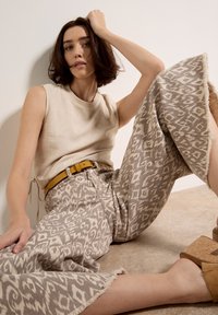 Donna seduta a terra che indossa un top beige senza maniche, pantaloni ampi con motivo e orlo sfrangiato, cintura gialla e sandali zeppa color cuoio.