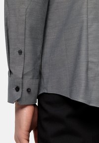 Camicia grigia a fantasia con tessuto texture, dotata di bottoni neri sui polsini e orlo dritto, abbinata a pantaloni scuri.