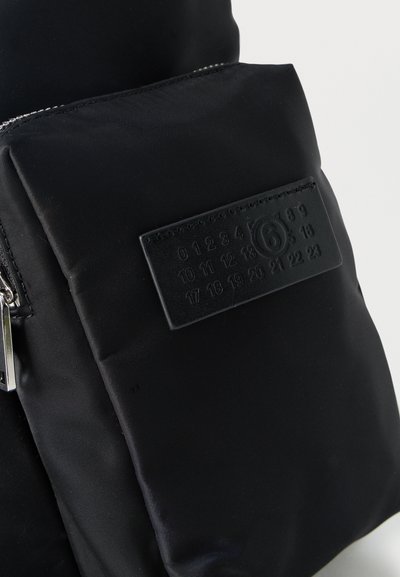 Μαύρη υφασμάτινη τσάντα με μπροστινή τσέπη με φερμουάρ και ραμμένη ορθογώνια ετικέτα που εμφανίζει ανάγλυφους αριθμούς από 0 έως 23, με τον αριθμό 6 κυκλωμένο.