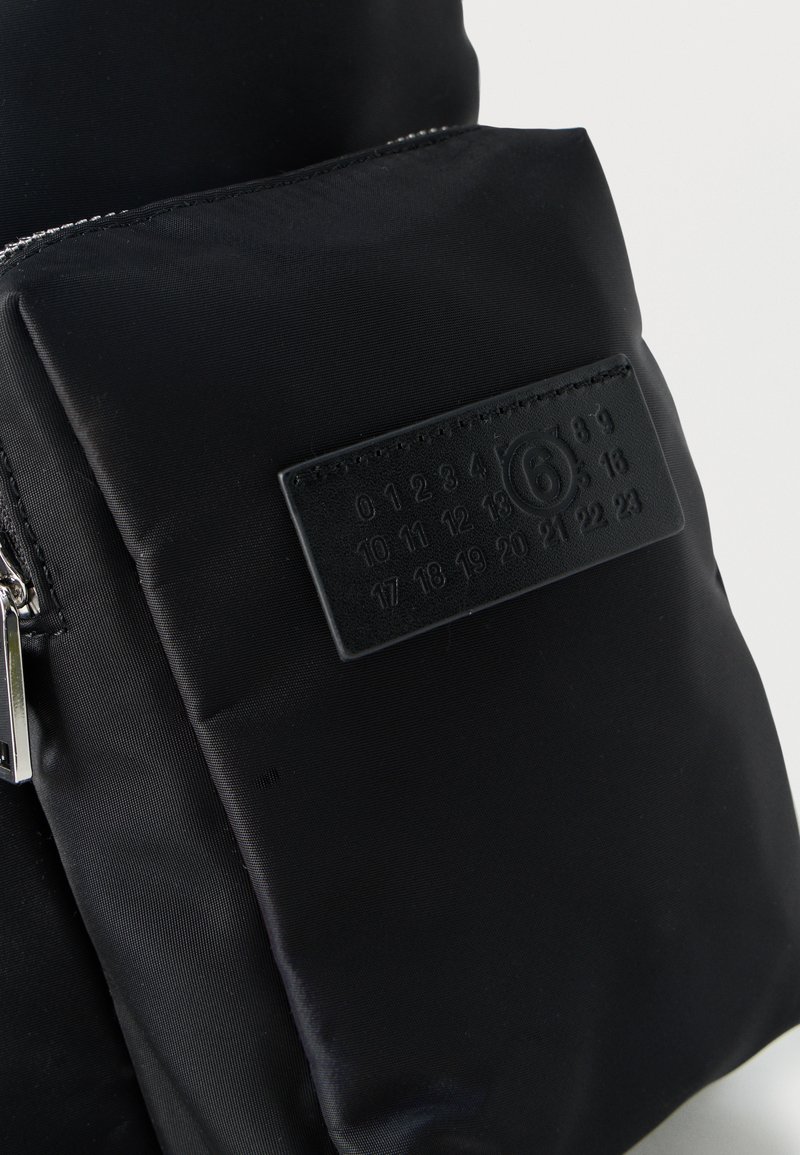 Sac en tissu noir avec poche à fermeture éclair à l'avant et patch rectangulaire cousu affichant des chiffres en relief de 0 à 23, avec le numéro 6 entouré.