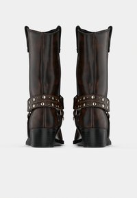 Σκούρα καφέ δερμάτινες μπότες μέχρι τη μέση της κνήμης με μαύρα τακούνια, μεταλλικά καρφιά σε λουριά γύρω από τους αστραγάλους, εμφανιζόμενες από πίσω σε λευκό φόντο.