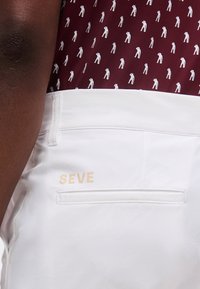 Κοντινό λευκού παντελονιού με το κείμενο "SEVE" πάνω από την πίσω τσέπη, φορεμένο με μπορντό πουκάμισο με μικρά λευκά σιλουέτες γκολφέρ.