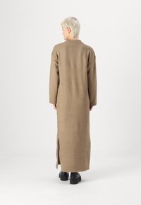Robe tricotée longue, marron, avec une silhouette droite, des fentes sur les côtés et un col côtelé. Texture douce et manches longues. Chaussures noires visibles.