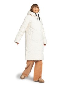 Manteau d'hiver blanc isolé avec capuche, présentant une texture matelassée, des poches avant et une fermeture éclair. Associé à un pantalon marron clair.
