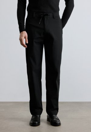 Pantalones negros con cintura ajustable mediante cordón, confeccionados en tela suave. El diseño es de corte recto, con bolsillos laterales y detalles mínimos.