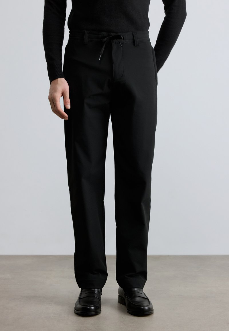 Čierne nohavice so sťahovacou šnúrkou v páse, vyrobené z hladkej tkaniny. Strih je rovný, s bočnými vreckami a minimálnymi detailmi.