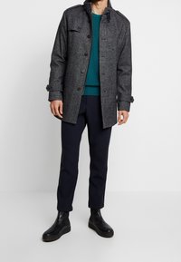 Grå ullblandad kappa med knappstängning och ståkrage, i kombination med en petrolblå stickad tröja och smala mörka byxor. Svarta ankelstövlar avslutar looken.