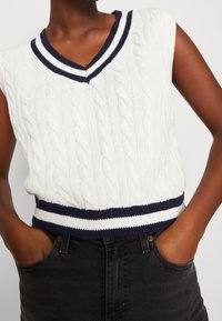 Bílý pletený svetr s copánkovým vzorem, námořnický pruhovaný výstřih do V a odpovídající lem. Nosený přes černé vysoké džíny.