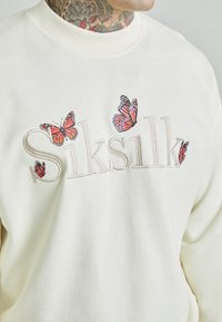 Krémová mikina se vyšitým nápisem "Siksilk" a designem oranžových a černých motýlů. Vyrobena z jemné tkaniny s hladkou texturou.