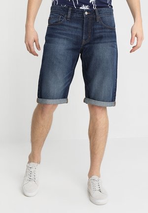 Mann trägt dunkelblaue, hochgekrempelte Jeansshorts und weiße Turnschuhe, steht vor einem schlichten hellen Hintergrund.