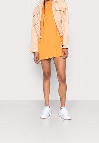 Kvinna i en ljus persikofärgad jacka, orange klänning med Nike-logotyp och vita sneakers, som står mot en helt ljusgrå bakgrund.