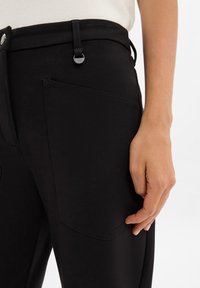 Black trousers with a smooth texture, featuring a side pocket and metal button detail. Waistband has adjustable straps for a tailored fit.