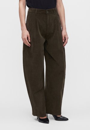 Femme portant un pantalon en velours côtelé marron foncé taille haute, à jambes larges avec des plis, des talons noirs à bout pointu et un haut noir, debout devant un fond uni.