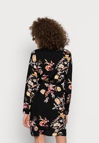 Donna con capelli ricci che indossa un vestito lungo a maniche lunghe nero con fiori, in piedi con la schiena rivolta verso uno sfondo chiaro e uniforme.