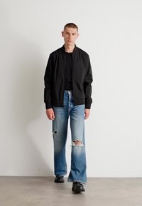 Black bomber jacket, black T-shirt, distressed light blue jeans with wide legs, and black boots. Model stands against a plain white background.