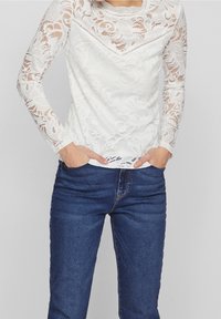 Top bianco in pizzo con maniche lunghe, motivo floreale e scollatura ondulata, indossato sopra jeans in denim blu. La texture sembra leggera e trasparente.