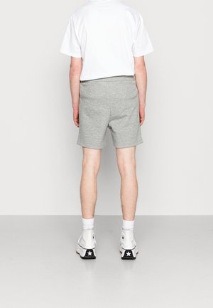Adulto con camiseta blanca, pantalones cortos grises, calcetines blancos y zapatillas blancas de caña alta, de pie con la espalda hacia el espectador sobre un fondo liso.