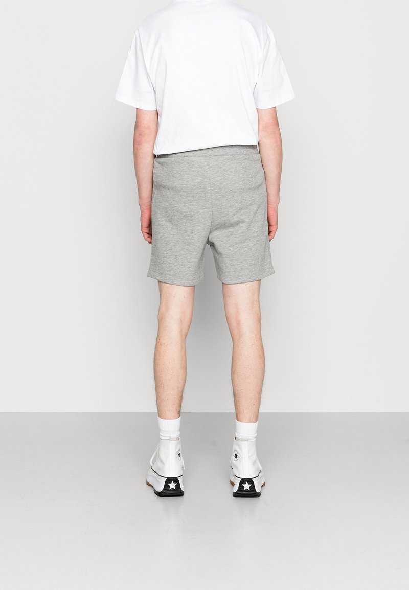 Adulte portant un t-shirt blanc, un short gris, des chaussettes blanches et des baskets montantes blanches, debout, dos à la vue sur un fond uni.