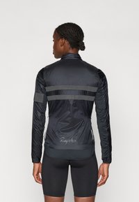 Sort cykeljakke med lange ærmer, reflekterende grå striber på tværs af ryggen og "Rapha" logoet ved kanten. Letvægts, strømlinet design.