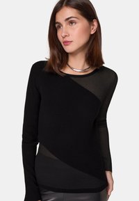 Schwarzer, gerippter Pullover mit langen Ärmeln und einem transparenten diagonalem Design. Verfügt über einen runden Halsausschnitt und eine glatte Textur, kombiniert mit einer metallischen Halskette.