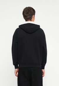 Sweatshirt preto com capuz, de textura suave, ombros caídos e cordão ajustável. Apresenta um bolso canguru e punhos canelados.