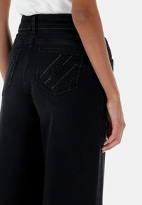 Persona che indossa jeans neri a vita alta con tasche posteriori decorate con strass e una camicia bianca infilata dentro, mostrata dalla vita in giù.