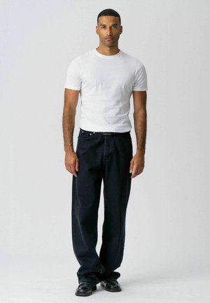 Mies seisoo neutraali ilme kasvoillaan, pukeutuneena istuvaan valkoiseen t-paitaan, löysiin mustiin farkkuihin ja mustiin kenkiin yksinkertaista harmaata taustaa vasten.