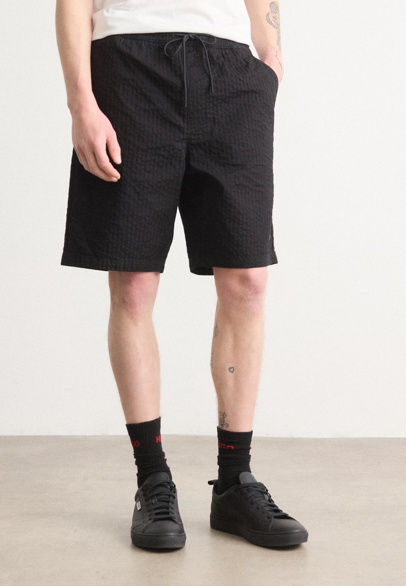 Schwarze gemusterte Shorts mit elastischem Bund und Kordelzug, ausgestattet mit seitlichen Taschen und einem lockeren Schnitt. Getragen mit schwarzen Schuhen und Socken.