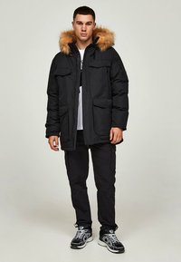 Svart dunjacka med pälsbeklädd luva, utrustad med två framfickor och dragkedja. Bärs med svarta cargobyxor och sportskor.