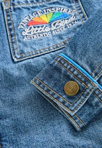 Jeans jakna z odprtnino s pisanem sončnim motivom in besedilom "Vintage Inspired". Zlati gumb z logotipom "Little Bird" in modrim notranjim obrobljenjem.