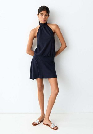 Jeune femme portant une robe sans manches bleu marine à col halter avec une jupe asymétrique et des sandales plates blanches, debout contre un mur blanc.