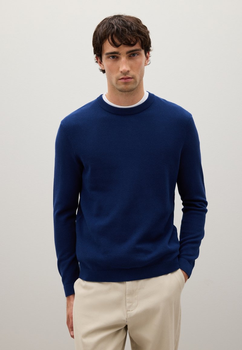 Maglione lavorato a maglia blu navy con colletto rotondo, maniche lunghe e polsini e orlo a costine. Indossato sopra una maglietta bianca, abbinato a pantaloni beige.