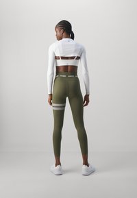 Vêtements de sport pour femmes : haut à manches longues blanc avec un dos transparent, leggings verts olive avec des bandes réfléchissantes et une ceinture "STRONGER". Chaussures blanches.