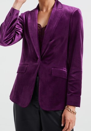 Žena v fialovém sametovém saku přes vršek s krajkovým lemováním, černých kalhotách a zlatém řetízkovém náramku na zápěstí.