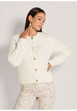 Femme portant un pull duveteux blanc boutonné et un pantalon clair à motifs, posant avec une main sur la hanche et l'autre près du cou.