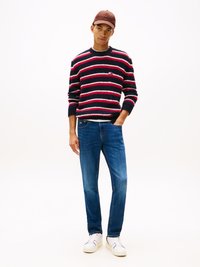 Tengerés kék és piros csíkos kötött pulóver, laza fazonban, kék farmernadrággal és fehér tornacipővel párosítva. Barna sapka teszi teljessé a megjelenést.