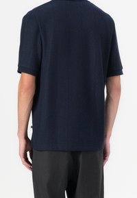 Homme portant une chemise texturée bleu marine foncé à manches courtes et un pantalon foncé à fines rayures, vu de dos sur fond blanc.