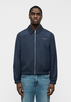 Άνδρας που φοράει μπλε ναυτικό μπουφάν με φερμουάρ και ελαστικές μανσέτες, με το λογότυπο Michael Kors, σε συνδυασμό με ανοιχτόχρωμο γαλάζιο τζιν, στέκεται μπροστά σε απλό φόντο.