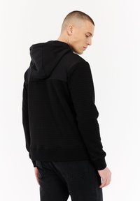 Jonge man met kort haar in een gestructureerde zwarte hoodie en zwarte jeans, die zijwaarts staat tegen een witte achtergrond.