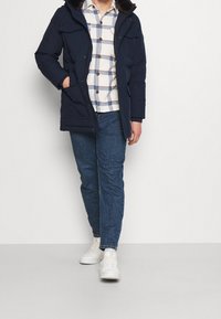 Hombre vestido con un abrigo parka azul marino, una camisa de cuadros blanca y azul, vaqueros azules y zapatillas blancas, de pie contra una pared blanca lisa.