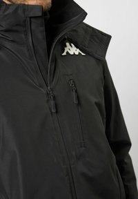 Schwarze Jacke mit hohem Kragen, ausgestattet mit zwei Reißverschlusstaschen und einem auffälligen weißen Markenlogo. Das Material wirkt glatt und matt.